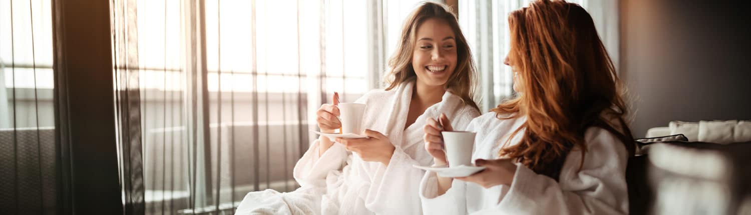 Zwei Frauen in Bademänteln trinken aus weißen Tassen auf einer Couch.